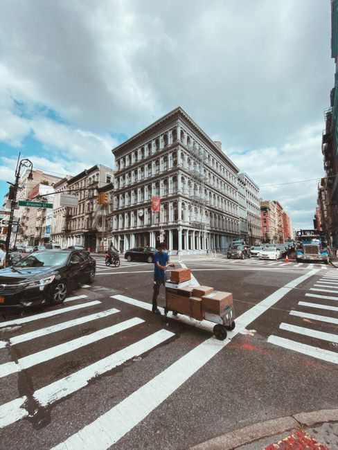 A street scene featuring a delivery worker pushing a hand truck loaded with several cardboard boxes across a pedestrian crossing in front of a historic, multi-story building with ornate architectural 