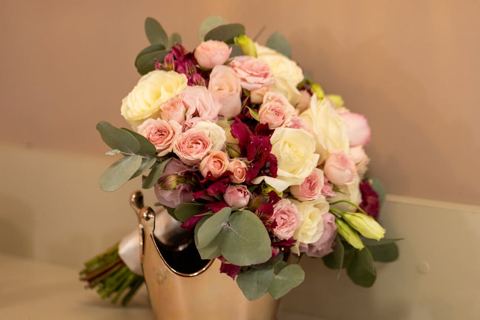 A bouquet of fresh flowers consisting of pale pink roses, creamy white roses, and deep burgundy blooms, accented with green eucalyptus leaves. The arrangement is tightly clustered, with the flowers ar