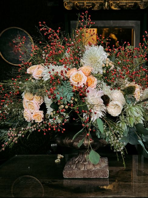 A lush floral arrangement featuring a variety of fresh flowers, including large white dahlias, pale peach roses, and smaller white blooms, complemented by red berries, green foliage, and sprigs of euc