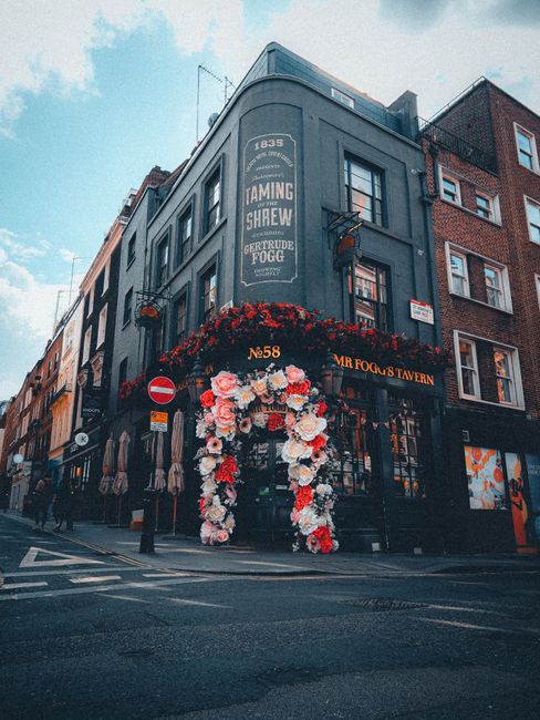 A woman with short, gray hair wearing a white cardigan is selecting a bouquet of fresh flowers at SoHo Flowers, located on Old Compton Street. She is reaching out with both hands to grasp a bouquet wr