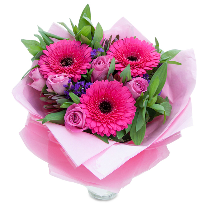 I like to think of this arrangement as a small, private celebration of pinks and purples, the kind of bouquet you might carry home after a detour past Dean Street or a browse at the nearby market. Three fuchsia gerbera daisies dominate the composition with their open, sun-like faces; their many petals fan out energetically around deep, dark centres that give the display a lively, contemporary edge. Nestled among them are roses in a softened lavender-pink or dusty rose, petals still gently curled inward like quiet promises, providing a romantic, tactile softness. Tiny sprays of purple statice are tucked throughout, lending a rustic texture and a cool counterpoint to the warmth of the pinks.
A generous layer of green foliage - broad, resilient leaves similar to salal and touches of eucalyptus - builds volume and adds a crisp, slightly aromatic undercurrent. The bouquet is lovingly wrapped: an inner layer of matte pale-pink tissue offers a muted base, while an outer sheet of sheer, slightly crinkled fuchsia-pink cellophane introduces a subtle shimmer and cohesion. The folds are deliberate and neat, reflecting a hands-on florist's attention, and the clear glass vase rim visible below suggests the flowers are meant to be displayed and enjoyed immediately.
In soft afternoon light this bouquet reads as both playful and composed, equally suited to brightening a bedside table or marking a significant anniversary. For anyone in Soho looking to offer warmth and celebration, this arrangement conveys affection, care and artisanal quality.